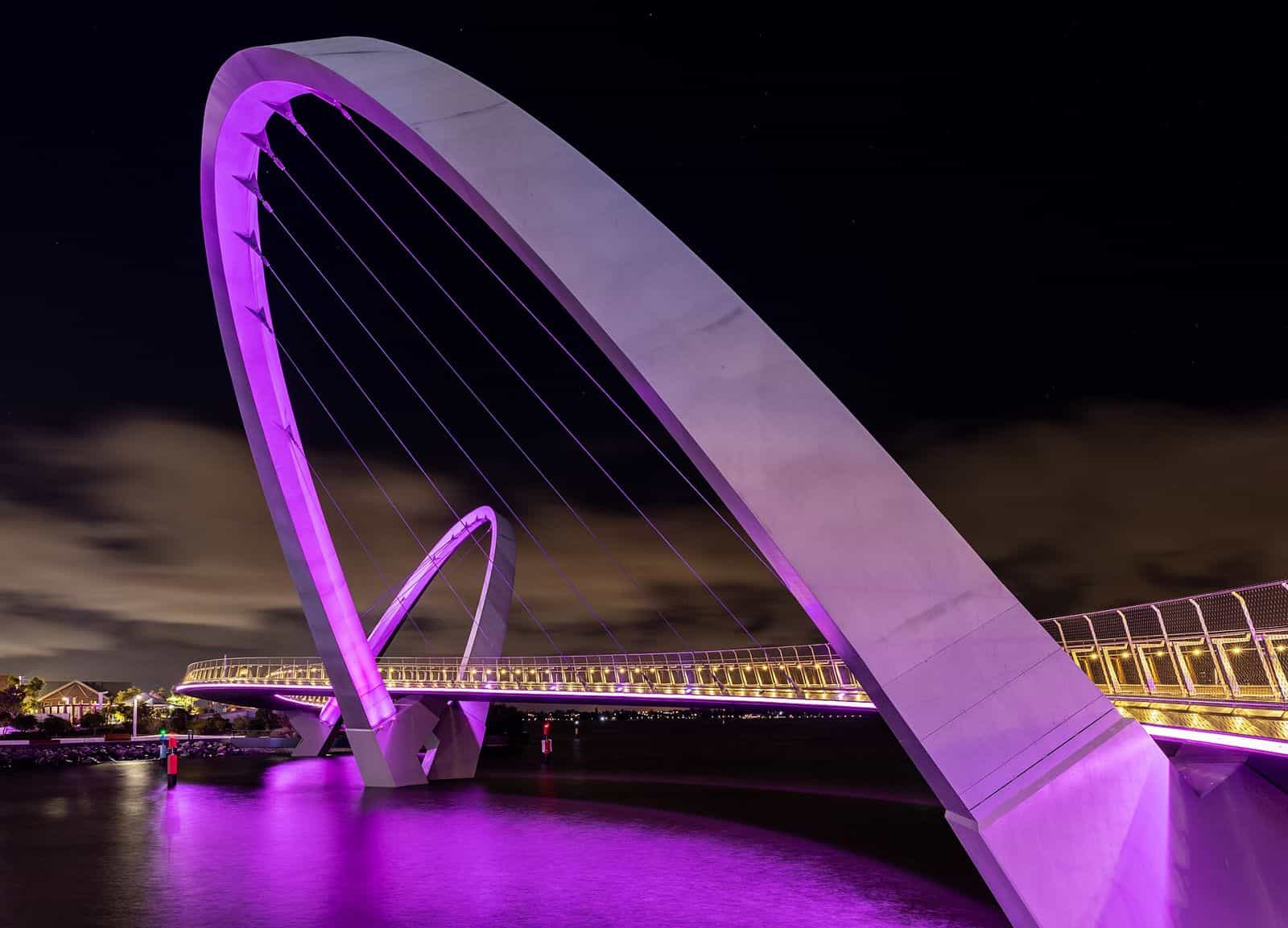 The curved Elizabeth Quay Bridge lit in purple at night, reflected in the water with city towers behind