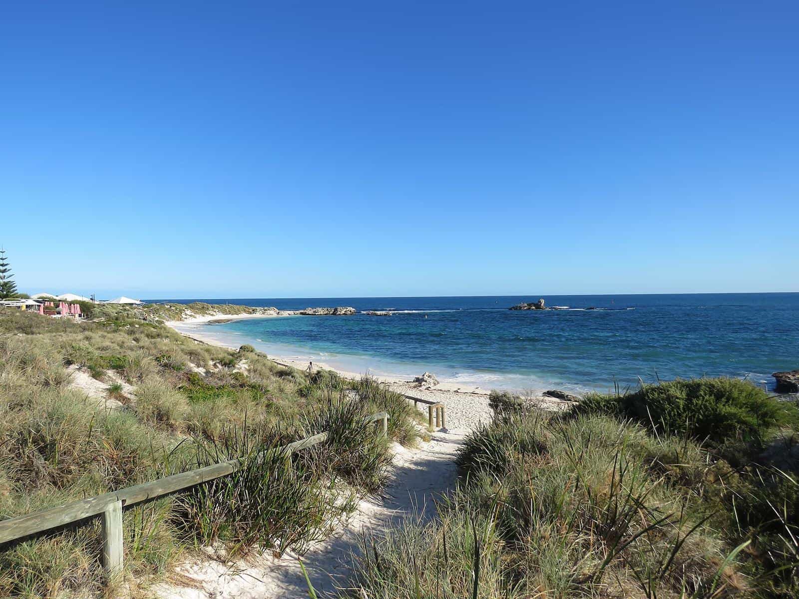 A sandy path leading down between low coastal shrubs to a turquoise bay on Rottnest Island
